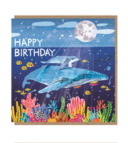 Square card with brown kraft envelope tucked inside. Illustration on the card is of an adult and baby blue whale swimming above a coral reef with different types of colourful fish. Above the water is a smiling moon, clouds and stars. The words 'happy birthday' are in white capital letters on either side of the water line.